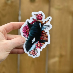 May include: A sticker featuring a black and white orca with pink and red flowers. The orca is swimming through the flowers, and the sticker has a white background.
