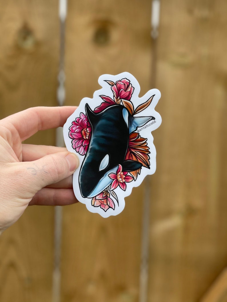 May include: A sticker featuring a black and white orca with pink and red flowers. The orca is swimming through the flowers, and the sticker has a white background.