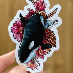 May include: A black and white orca whale with a white eye patch is surrounded by pink and red flowers. The orca is facing the left side of the image. The background is white. The text "@amttattoos66" is in the upper right corner of the image.