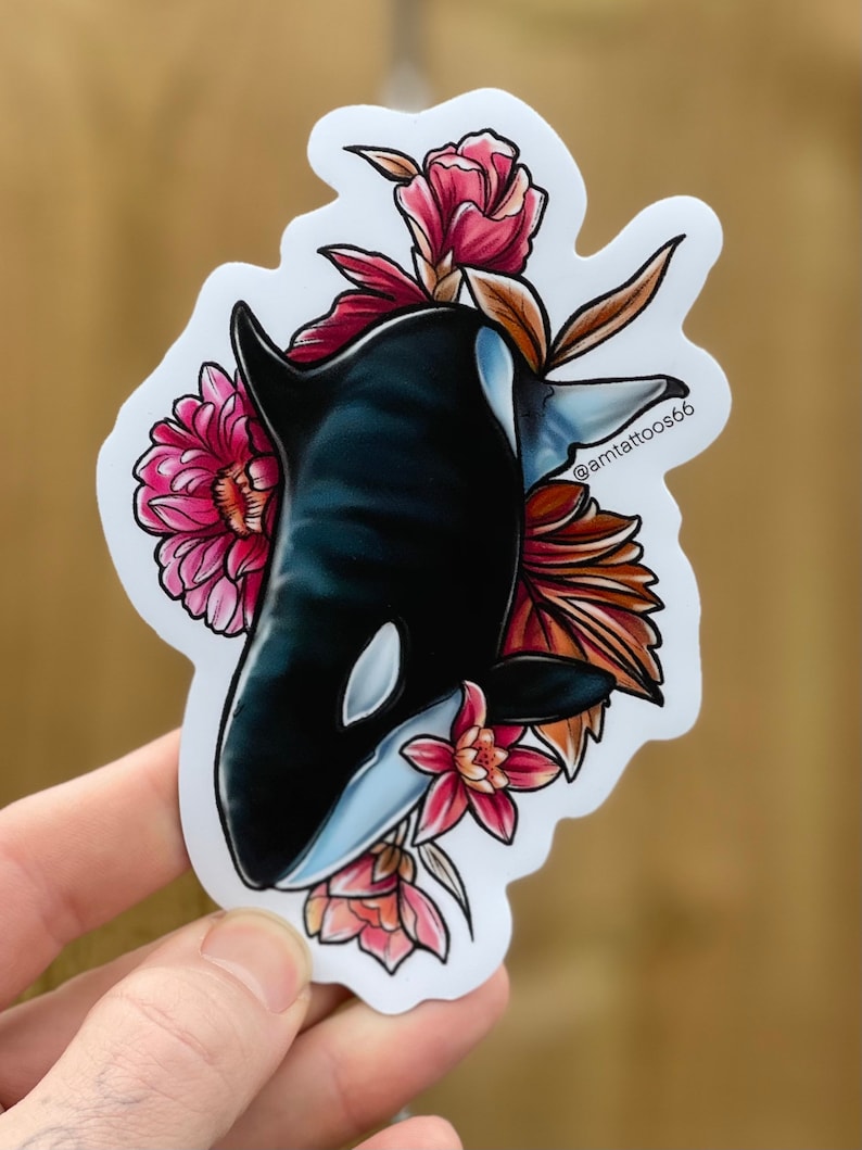 May include: A black and white orca whale with a white eye patch is surrounded by pink and red flowers. The orca is facing the left side of the image. The background is white. The text "@amttattoos66" is in the upper right corner of the image.