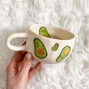 Könnte beinhalten: Cremefarbene Keramiktasse mit großem Henkel, verziert mit grünen Avocado-Illustrationen. Die Avocados haben lächelnde Gesichter und orangefarbene Details. Die Tasse steht auf einer weißen, flauschigen Oberfläche.