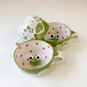 Könnte beinhalten: Drei Keramikbecher mit einem skurrilen Design. Die Becher sind weiß mit grünen Akzenten und zeigen ein Froschgesicht im Inneren. Die Becher haben rote Tupfen und Pilzillustrationen. Jeder Becher hat einen Henkel.