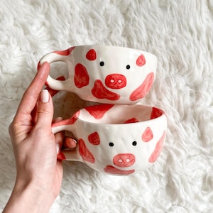 Könnte beinhalten: Zwei weiße Keramikbecher mit roten kuhartigen Flecken und einem Schweinegesicht-Design. Jeder Becher hat einen Henkel und schwarze Punktaugen. Die Becher sind übereinander gestapelt.