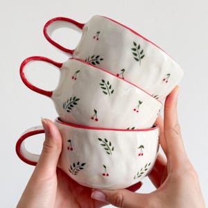 Może przedstawiać: Stos trzech białych ceramicznych kubków o falistej, teksturowanej powierzchni. Każdy kubek posiada ręcznie malowany wzór w małe czerwone wiśnie i zielone liście. Krawędzie oraz uszka kubków są pomalowane na jednolity czerwony kolor.