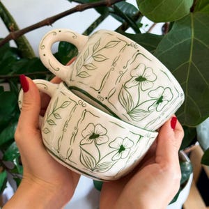 Op de afbeelding: Twee witte keramische mokken op elkaar gestapeld, elk versierd met een groen bloemmotief. De mokken hebben een afgeronde vorm met een handvat en zijn versierd met groene bladeren en bloemen. De mokken worden voor een groene, bladerrijke achtergrond gehouden.