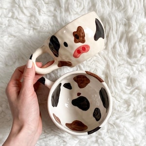 Può includere: Un set di tazza e ciotola in ceramica con un design a mucca. I pezzi bianco sporco presentano macchie marroni e nere, con la tazza che mostra una faccia di mucca dipinta con narici rosse. La ciotola ha un motivo simile, creando un set stravagante a tema fattoria.