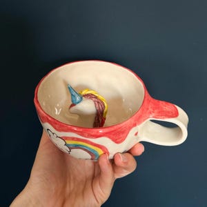 Könnte beinhalten: Eine weiße Keramiktasse mit rotem Rand und Henkel, mit einem 3D-Einhornkopf im Inneren. Die Tasse hat ein Regenbogen- und Wolken-Design an der Seite. Das Einhorn hat ein blaues Horn, eine gelbe Mähne und einen rosa Schwanz.