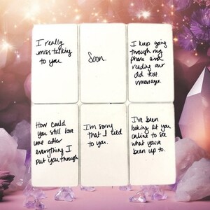 May include: Six white cards with handwritten text, including phrases like "I really miss talking to you" and "I'm sorry that I lied to you." The cards are arranged on a pink surface with crystals and a soft, blurred background.
