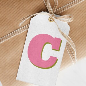 May include: A wrapped gift in brown paper, tied with twine. A white tag with a pink letter "C" and green outline hangs from the twine. The background is a white surface.