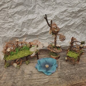 May include: A miniature fairy garden set featuring a bench, chair, and tiered display stand, all crafted from twigs and twine. The set includes a blue flower and is decorated with moss and small white flowers. The items are arranged on a wooden surface.