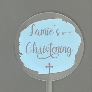 Può includere: Topper per torta in acrilico trasparente con la scritta "Jamie's Christening" in un carattere corsivo, su uno sfondo azzurro chiaro in stile acquerello. Una croce in basso. Il topper ha un bastoncino appuntito per l'inserimento.