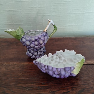 Può includere: Un set composto da lattiera e zuccheriera in ceramica a forma di grappoli d'uva viola. La lattiera presenta un beccuccio a forma di foglia verde e un manico curvo, mentre la zuccheriera aperta ha un esterno testurizzato e un interno bianco.