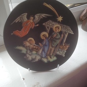 Può includere: Un piatto in ceramica nera con un'immagine in rilievo della scena della Natività. La scena include una stella, un angelo con le ali, una mangiatoia, una mucca, una pecora e tre figure. Le figure sono una donna con una veste blu, un uomo con una veste marrone e un uomo con una veste rossa.