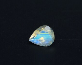 May include: A pear-shaped, faceted gemstone with a rainbow iridescence. The gem displays a play of colours, including blues, yellows, and greens, against a dark background. The gemstone is cut to maximise light reflection.