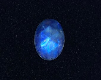 May include: A blue moonstone cabochon gemstone with a faceted cut. The stone is oval shaped and has a blue iridescent sheen.