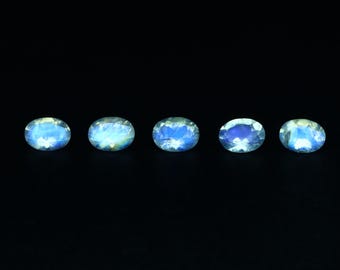 May include: Five oval-shaped, iridescent gemstones with a blue and white color scheme are arranged in a horizontal line against a black background. The gemstones appear to be polished and reflect light, showcasing their internal structure.