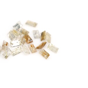 May include: A collection of small, rectangular gemstones with a mix of clear, light brown, and golden hues. The gemstones are scattered against a white background, showcasing their varied colors and internal structures. The stones appear to be cut with facets.