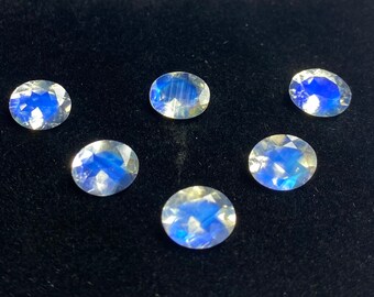 May include: Six oval-shaped moonstone gemstones with a blue iridescent sheen. The stones are arranged in a semi-circle on a black surface.