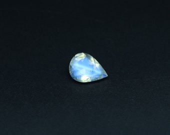 May include: A pear-shaped moonstone gemstone with a blue iridescent sheen. The stone is set against a black background.