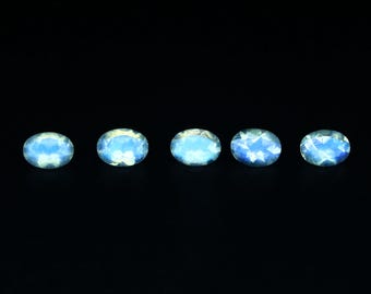 May include: Five oval-shaped, iridescent gemstones are displayed against a black background. The stones have a pale blue and white color, with a subtle shimmer. The gemstones are arranged in a horizontal line.