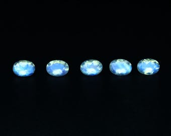 May include: Five oval-shaped, iridescent gemstones with a blue and white color scheme are arranged horizontally against a black background. The gemstones have a faceted surface, catching the light and creating a shimmering effect. These gemstones could be used for jewelry making.