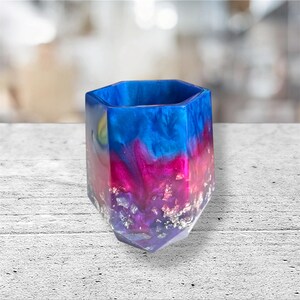 May include: A hexagonal resin container in shades of blue, pink, and purple, with silver leaf detailing. The top is blue, fading into pink and purple towards the base. The container has a geometric design.