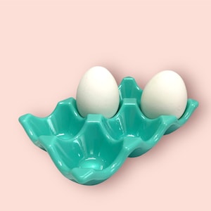 May include: A turquoise ceramic egg holder containing two white eggs. The holder has a wavy design and is set against a light pink background. The eggs are oval-shaped. The holder is approximately 15 cm long.