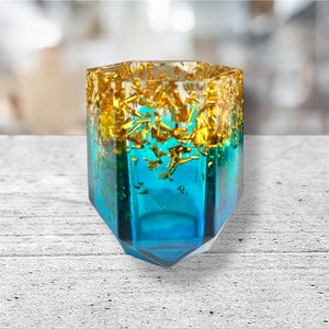 May include: A hexagonal teal and gold resin votive candle holder. The top edge is embedded with gold flakes, transitioning to a clear teal body. The geometric design adds a modern touch to home decor.
