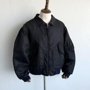 Könnte beinhalten: Eine schwarze Bomberjacke mit klassischem Kragen, durchgehendem Frontreißverschluss und zwei schrägen Vordertaschen. Die Jacke verfügt über eine kleine Reißverschlusstasche am linken Ärmel sowie dunkelblaue Rippbündchen an Ärmeln und Saum.