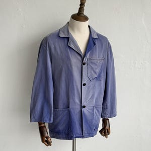 May include: A faded blue work jacket featuring a collared neckline, three dark buttons down the front, one chest pocket, and two lower pockets. The jacket has long sleeves and a slightly worn, textured appearance, displayed on a mannequin.
