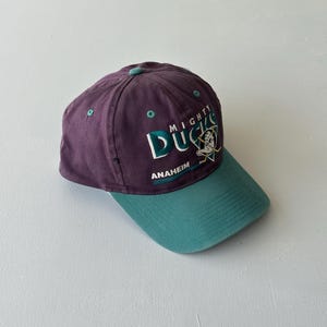 Può includere: Un cappellino da baseball viola con visiera verde acqua e un bottone verde acqua sulla parte superiore. La parte anteriore presenta un logo ricamato dei Mighty Ducks con una maschera da portiere, bastoni da hockey incrociati e il testo.
