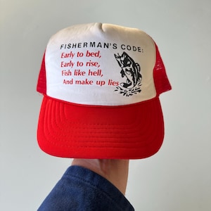 May include: A red and white trucker hat featuring the text "FISHERMAN'S CODE: Early to bed, Early to rise, Fish like hell, And make up lies" and a black fish illustration. The hat has a red peak and mesh back.