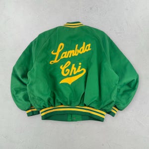 Könnte beinhalten: Eine grüne Collegejacke mit dem in gelber Schreibschrift gestickten Text Lambda Chi auf der Rückseite. Die Jacke verfügt über einen gerippten Kragen, Bündchen und Saum mit gelben und grünen Streifen, lange Ärmel und eine Knopfleiste.