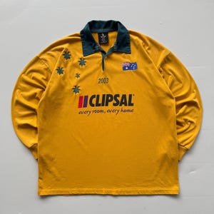 Könnte beinhalten: Senfgelbes Langarmshirt mit dunkelgrünem Kragen und Viertelreißverschluss. Mit dem Text "CLIPSAL" und "every room, every home" in Rot und Weiß. Enthält die australische Flagge und das Jahr "2003".