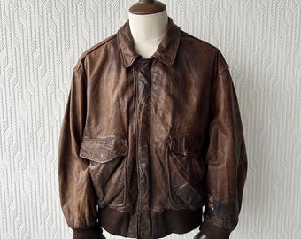 May include: A brown leather bomber jacket with a classic design, featuring a collared neckline, two front pockets, and ribbed cuffs and hem. The jacket is displayed on a mannequin, showcasing its vintage style and distressed texture. A small flag is visible on the sleeve.
