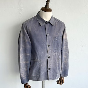 May include: A faded blue work jacket featuring a pointed collar, a front button closure, and three patch pockets. The fabric displays a distressed, mottled wash with light-colored paint splatters across the front, sleeves, and pocket areas.