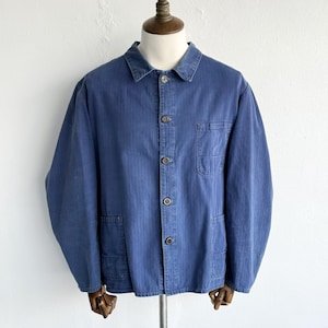 May include: A blue long-sleeve jacket featuring a herringbone weave pattern, a standard collar, and a front button closure. The garment includes one chest pocket on the left side and two lower patch pockets, all constructed from a durable cotton fabric.