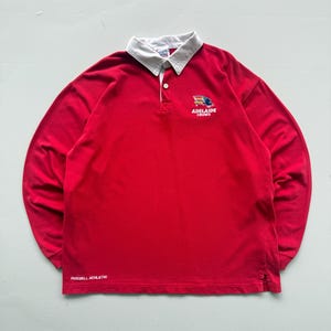 Könnte beinhalten: Ein rotes Rugby-Langarmshirt mit weißem Kragen. Das Shirt zeigt den Text "ADELAIDE CROWS" und "RUSSELL ATHLETIC". Es hat eine Knopfleiste mit drei Knöpfen und ein Logo auf der Brust.