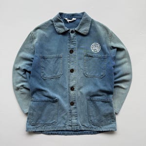 Op de afbeelding: Een verbleekte blauwe spijkerjas met vier voorzakken en een knoopsluiting. De jas heeft een klein wit logo op de borst en een klassieke kraag. De mouwen hebben een gradiënteffect, van donkerder naar lichtere blauwtinten.