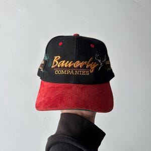 Könnte beinhalten: Eine schwarze Baseballkappe mit einem roten, wildlederartigen Schirm. Auf der Vorderseite befindet sich der gestickte Schriftzug Bauerly COMPANIES in Gold und Orange, ergänzt durch ein gesticktes Hirschkopfmotiv und kleine grüne Bäume.