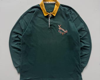 Puede incluir: Una camiseta de rugby verde oscuro de manga larga con cuello amarillo. La camisa presenta un diseño bordado en el pecho. El cuello tiene el texto "Line Out" impreso.