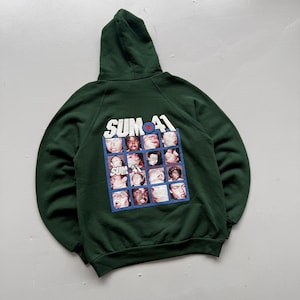 May include: A dark green hooded sweatshirt featuring a graphic print on the back. The print displays the text "SUM 41" above a grid of sixteen individual facial portraits of various people, with a red star inside a blue circle replacing the letter O.