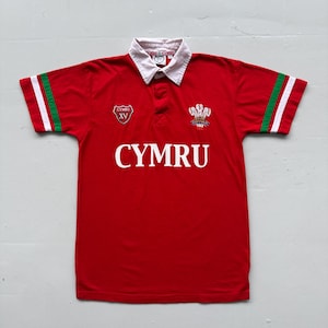 Peut inclure: Maillot de rugby rouge avec col blanc et manches rayées vertes et blanches. Le mot "CYMRU" est imprimé en grandes lettres blanches sur la poitrine. Comprend un écusson et le texte "CYMRU XV".