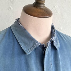 以下が含まれることがあります： 色あせたブルーのワークジャケット。クラシックな襟、ボタン付きカフスの長袖、フロントに3つのパッチポケットが付いています。前立てに沿った軽いペンキの飛沫や小さな補修パッチなど、着用による使用感が見られるデザインです。