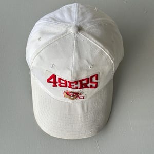 Op de afbeelding: Een witte honkbalpet met de rood geborduurde tekst 49ERS in een boog boven een klein rood en goudkleurig footballhelm-logo. De pet heeft een gestructureerde kroon, een gebogen klep en een knoopje aan de bovenkant voor een strak ontwerp.