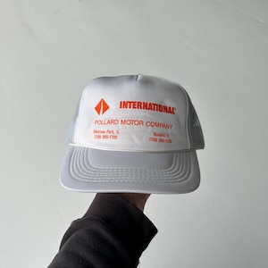 Pode incluir: Um boné trucker cinza claro com parte traseira em rede e painel frontal sólido. A frente apresenta um logótipo laranja com o texto INTERNATIONAL, POLLARD MOTOR COMPANY, Melrose Park, IL (708) 865-1700 e Roselle, IL (708) 980-6300 em laranja.