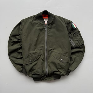May include: An olive green bomber jacket featuring a front zip fastening, ribbed collar, cuffs, and hem. It includes two front flap pockets with press studs and a utility pocket on the left sleeve adorned with a circular Italian flag patch.