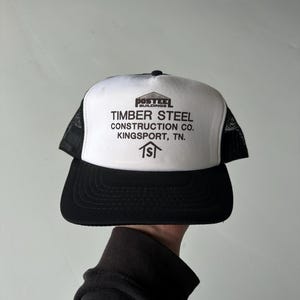 May include: A black and white trucker cap featuring a white front panel with black text that reads POSTEE BUILDINGS TIMBER STEEL CONSTRUCTION CO. KINGSPORT, TN. and a small house icon. The cap has a black brim and black mesh side and back panels.