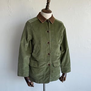 May include: An olive green corduroy jacket featuring a contrasting brown corduroy collar, long sleeves, and a front button closure. The garment includes two lower patch pockets and a vertical seam construction, displayed on a mannequin torso.