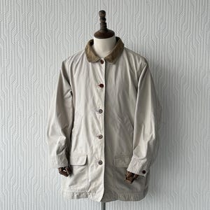 Puede incluir: Chaqueta beige con botones y cuello de pana marrón. La chaqueta tiene dos bolsillos delanteros y botones marrones. Se muestra en un maniquí con cabeza y brazos de madera.